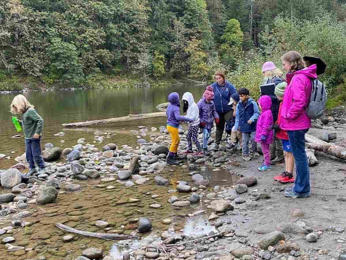 Group of students with teachers outdoors by a stream in the pine wooded background for article Franciscan Montessori Earth School for newcomers moving to Portland.