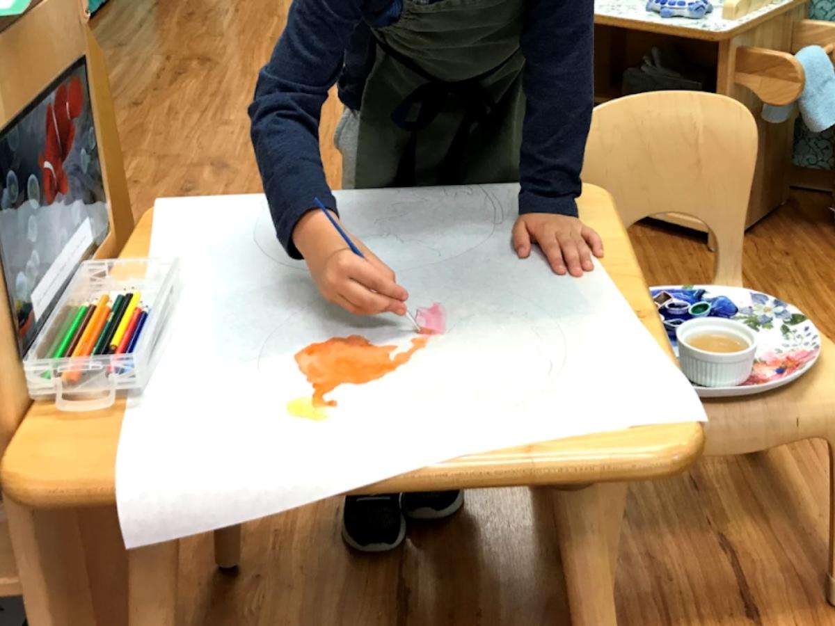 Child standing at a desk water coloring for article West Hills Montessori Elementary for newcomers moving to Portland.