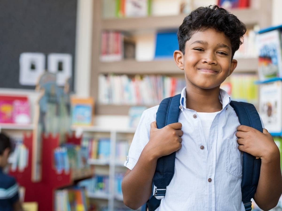 Boy smiling with a school backpack on standing in a classroom with bookshelves filled with books in background for article Estacada School District for newcomers moving to Portland.