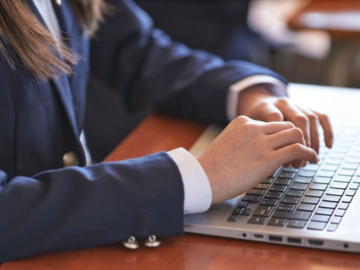 Close up of student in blue blazer with arms resting on desk and hands on laptop for article Valley Catholic School for newcomers moving to Portland.