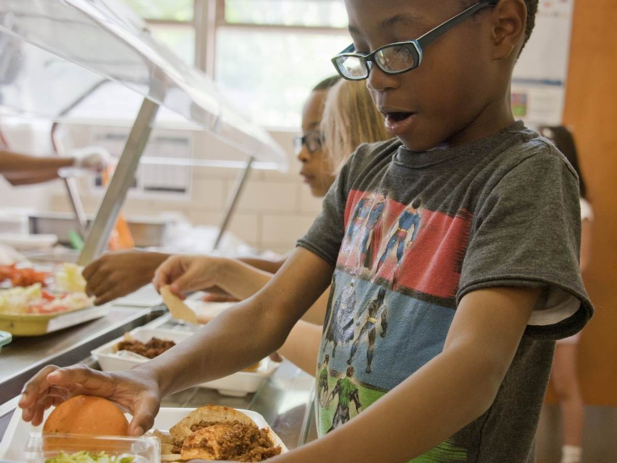 children in school lunch line getting their food with boy closest to camera looking with amazement at the food he is putting on his tray for article Portland Public Schools for newcomers moving to Portland area.