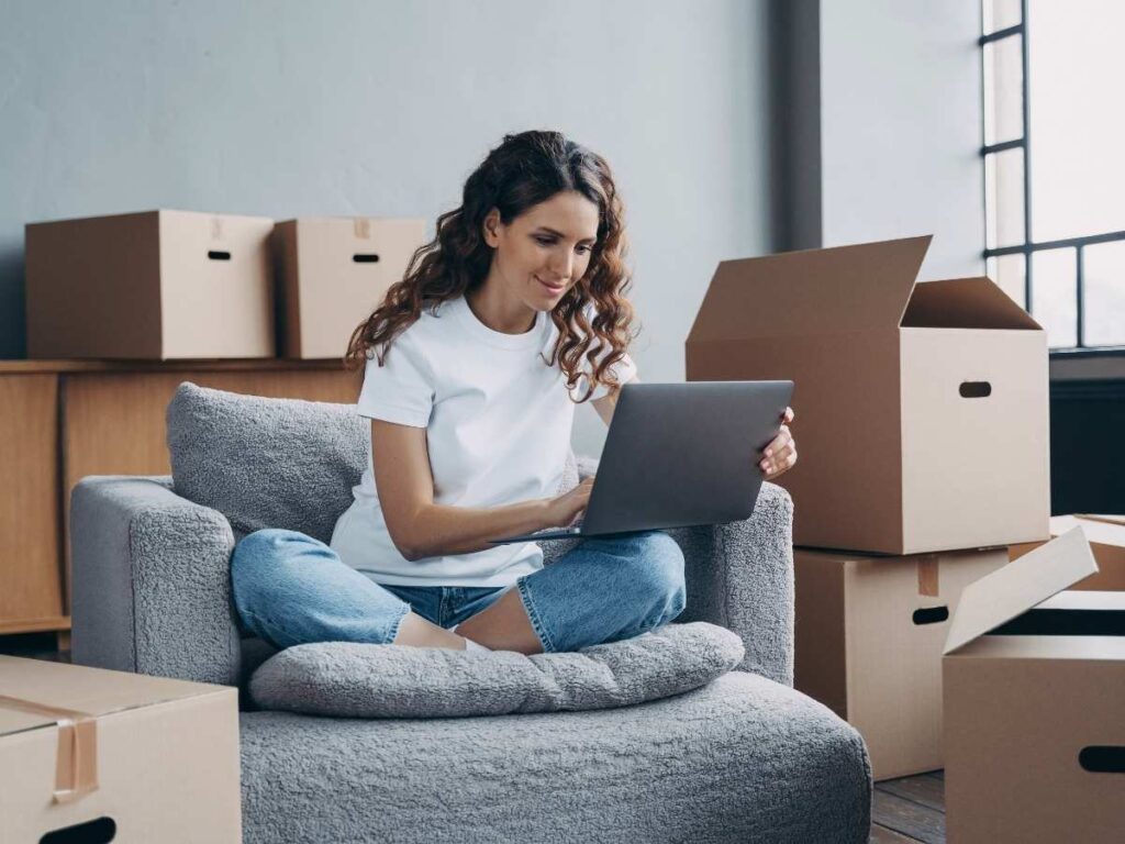 female sitting on chair with her legs crossed and laptop open on her lap in apartment with boxes stacked all around her for article Tenants Rights in Portland for newcomers moving to Portland.