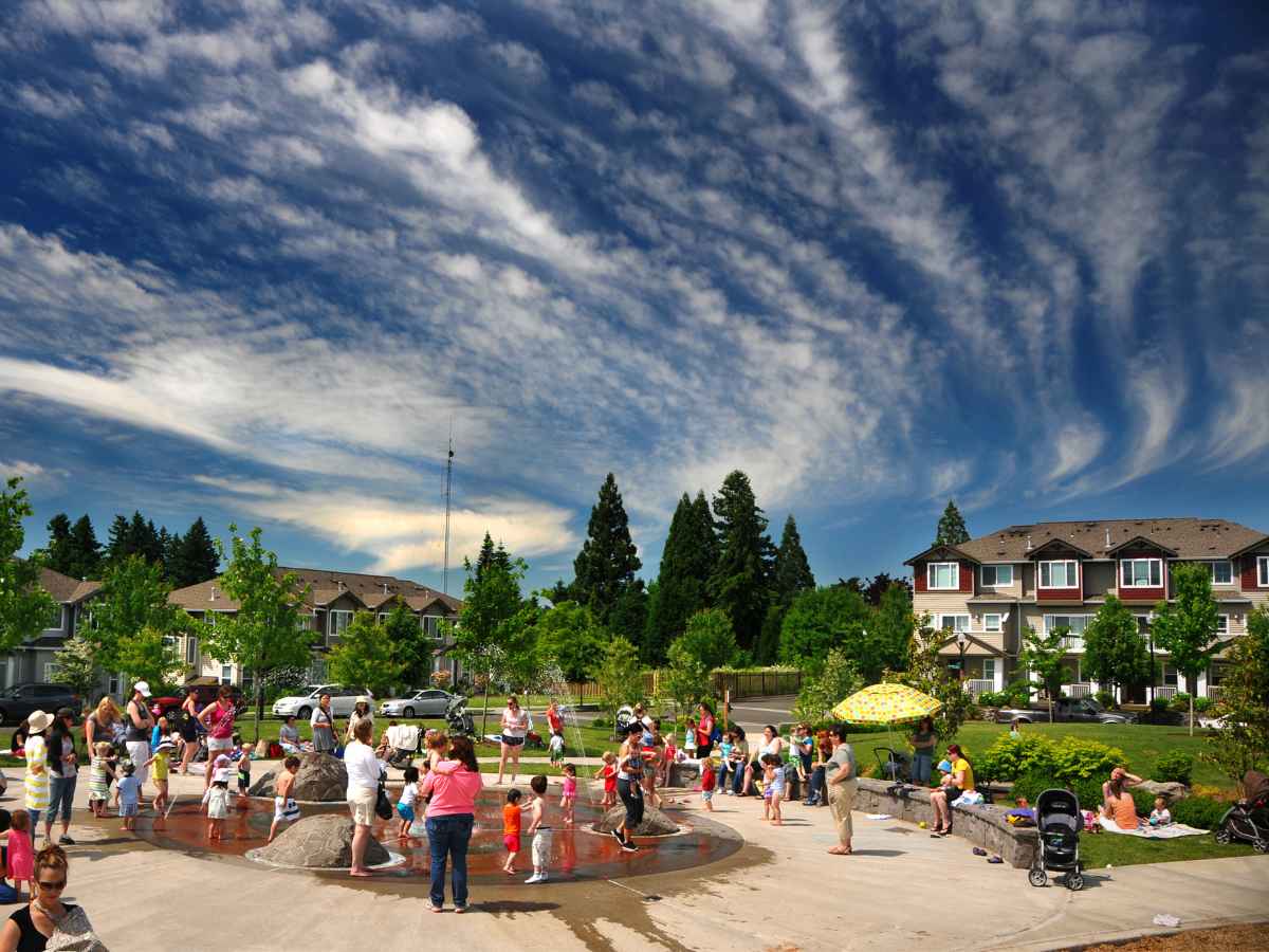 Magnolia park with people around a water park and townhomes in the background. Blue sky with white swirling clouds. For article Hillsboro, Oregon for newcomers moving to Portland.