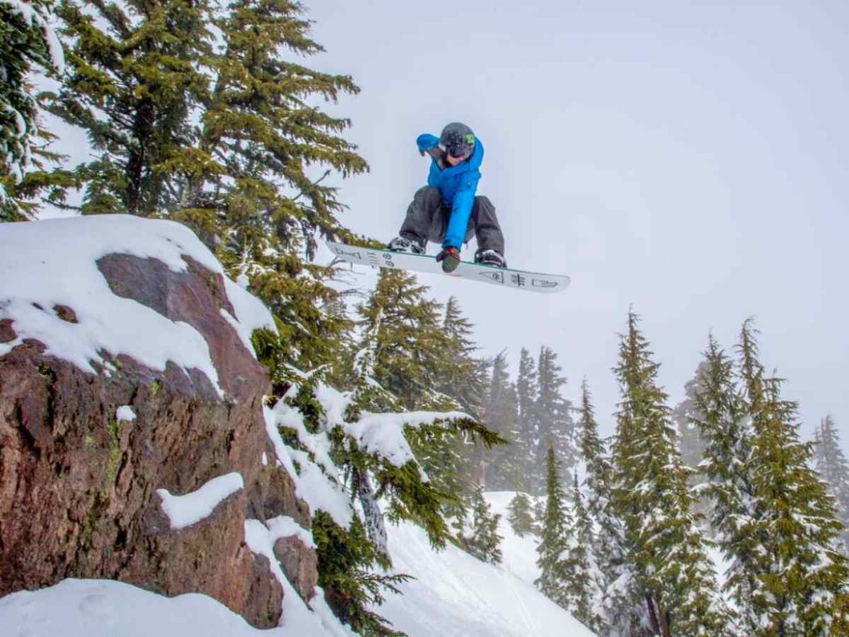 Snowboarder in blue and black snow gear catching air and holding board with one hand while going down snowy mountain surrounded by large rock formations and snow covered pine trees for article Skiing and Snowboarding Near Portland for newcomers moving to Portland.