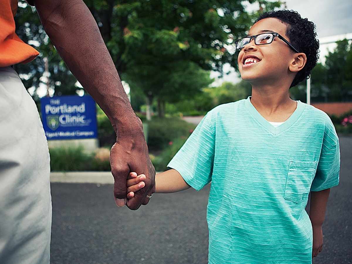 Image of a child with glasses in a green shirt holding hand of adult and looking up smiling at adult while walking outdoors for article The Portland Clinic for newcomers moving to Portland.