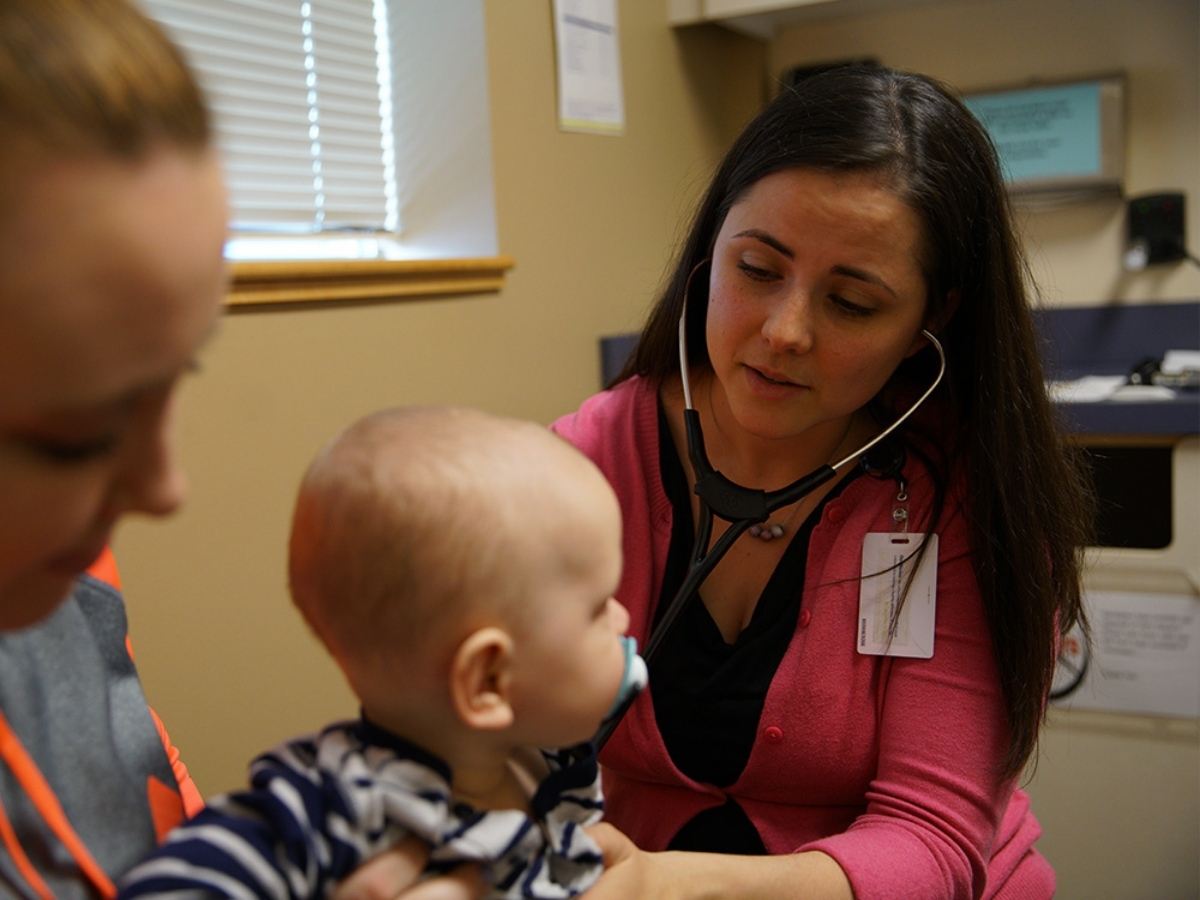 Mother holding baby while doctor examines baby with stethoscope for article The Portland Clinic - Treating People Like Family for newcomers moving to Portland.