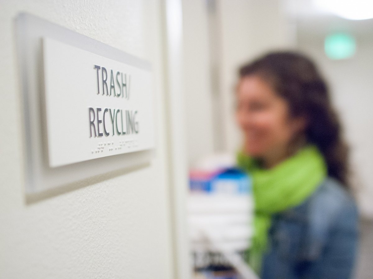 Close up of sign on side of office door that says Trash Recycle with a blurred woman's image standing in the doorway for article