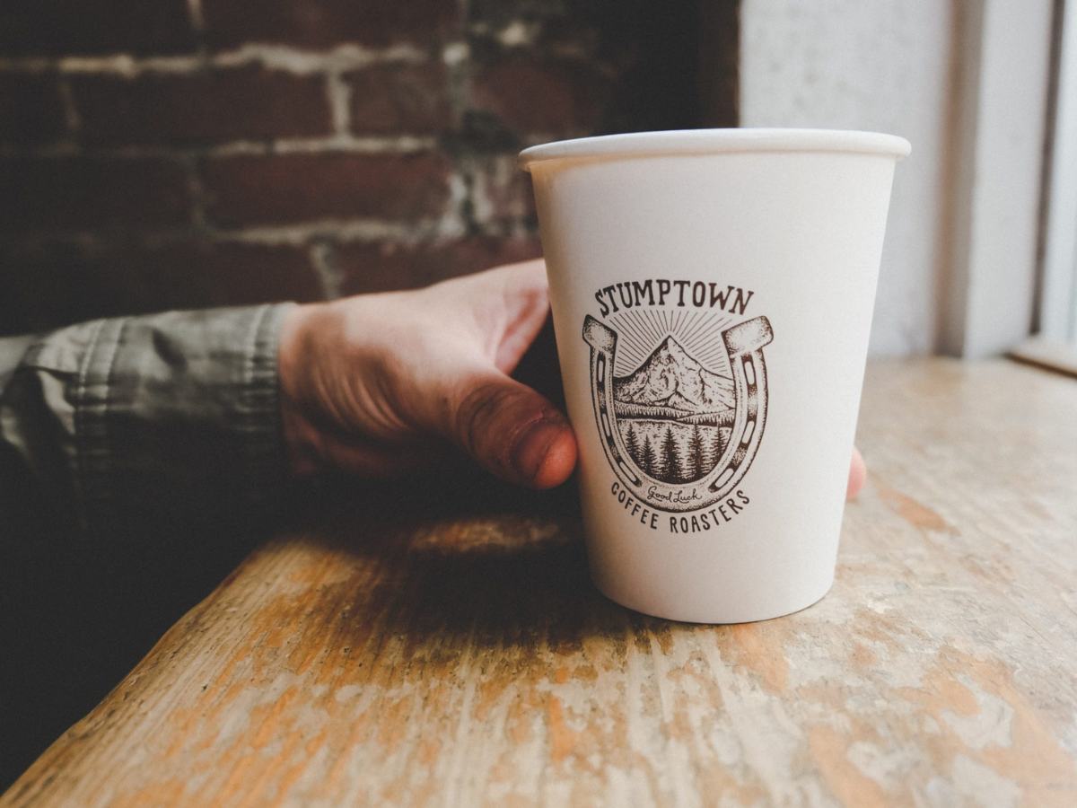 Close up of a hand clasped around a paper coffee cup that says Stumptown on it with a horseshoe as it sits on a wood table ledge and a brick background for article Best Communities for Families in Portland for newcomers moving to Portland.