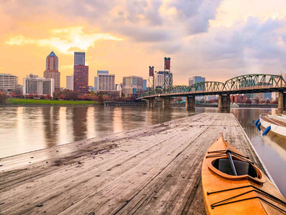 Downtown Portland, Oregon Panorama with Hawthorne Bridge. Sunset scene with dramatic sky and light reflections on the Willamette River. A dock and kayak in the foreground for article Cascade Sotheby’s International Realty for newcomers moving to Portland.