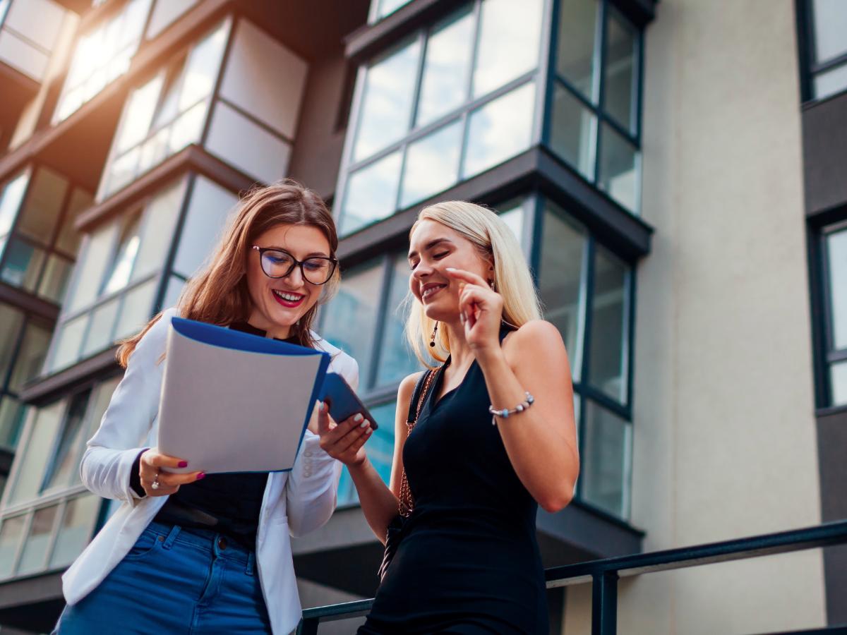 Realtor and client looking at list with modern apartment building behind them for article Renting vs Buying in Portland for newcomers moving to Portland.