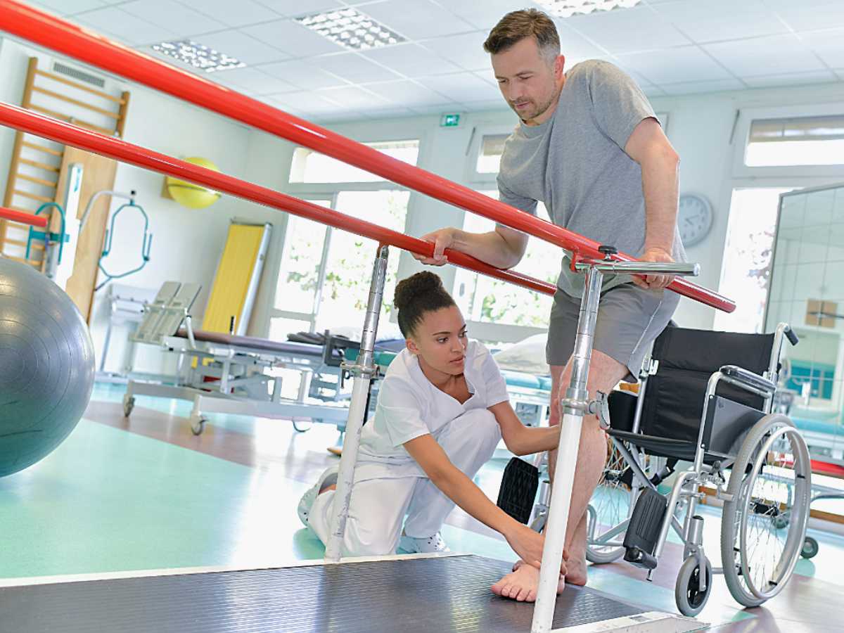 Man standing out of wheelchair on bars to help relearn to walk while physical therapist guides his feet for article Portland Physical Therapy: 6 Top-Rated Clinics for newcomers moving to Portland.