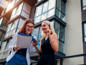 Realtor and client looking at list with modern apartment building behind them.