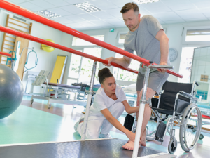 Man standing out of wheelchair on bars to help relearn to walk while physical therapist guides his feet.